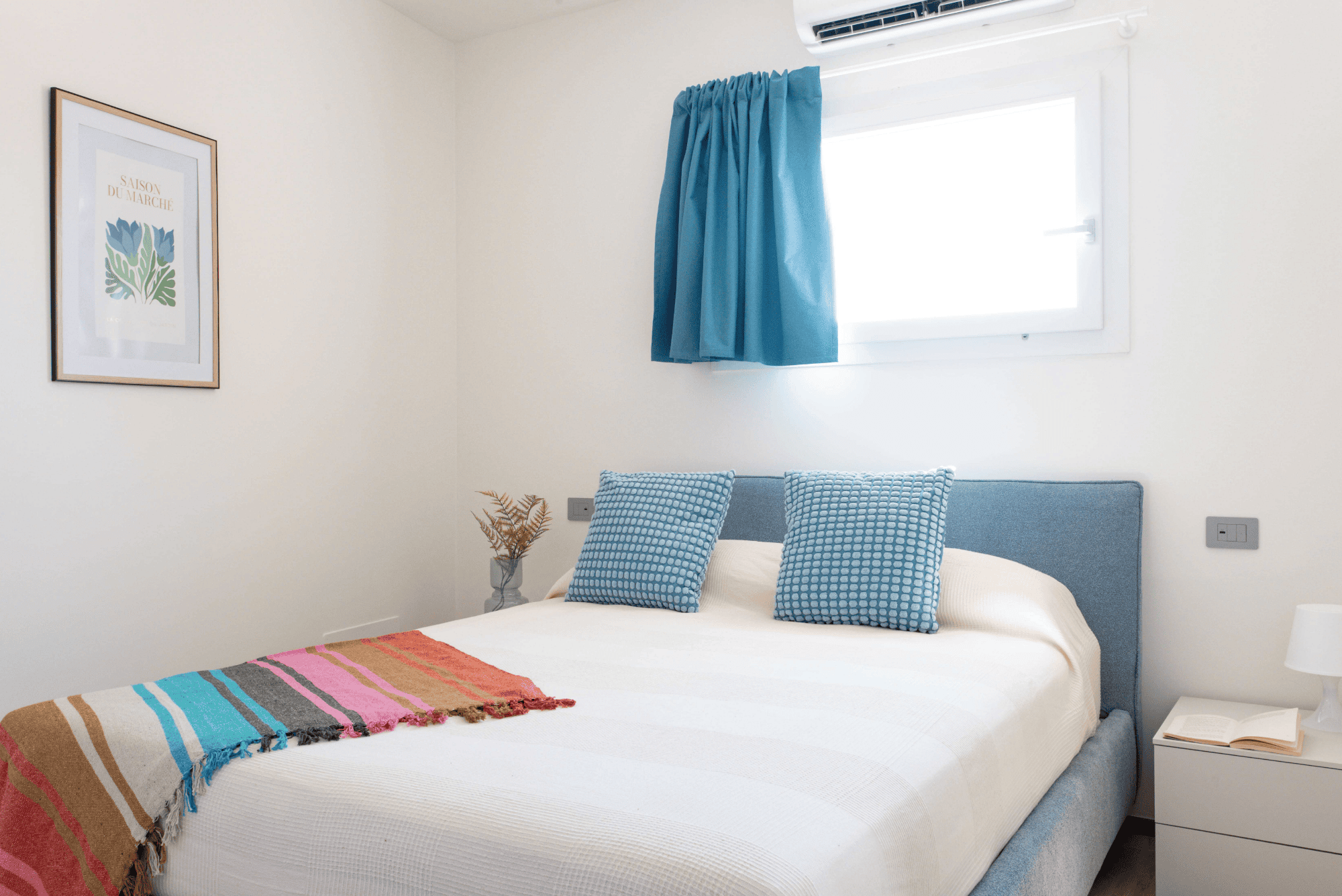 La camera da letto matrimoniale luminosa con finestra e tende e cuscini blu con una coperta multicolor sul letto