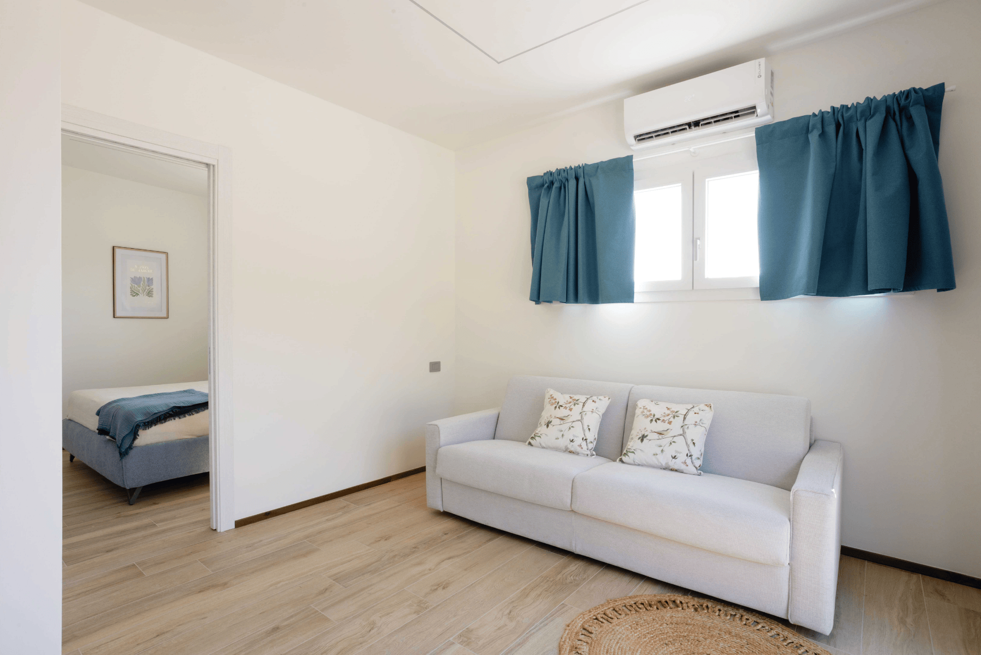La camera con divano letto grigio chiaro, sormontato da una finestra con tende blu