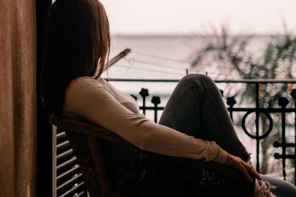 A woman from behind sitting on a small terrace facing the sea, looks towards the horizon.